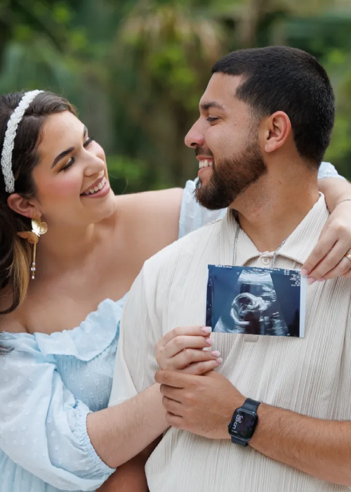 A couple joyfully holds an ultrasound image outdoors, celebrating their upcoming arrival, surrounded by greenery. A couple joyfully holds an ultrasound image outdoors, celebrating their upcoming arrival, surrounded by greenery.