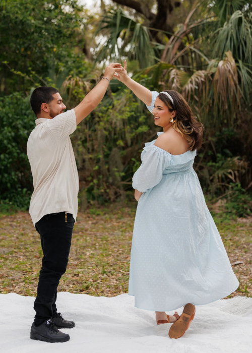A couple joyfully dances together outdoors, surrounded by lush greenery, with the woman in a light blue dress and the man in a casual outfit. A couple joyfully dances together outdoors, surrounded by lush greenery, with the woman in a light blue dress and the man in a casual outfit