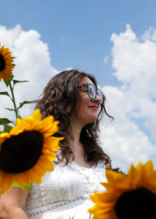 A woman stands amidst vibrant sunflowers, with a bright blue sky and fluffy clouds in the background, radiating summer joy A woman stands amidst vibrant sunflowers, with a bright blue sky and fluffy clouds in the background, radiating summer joy