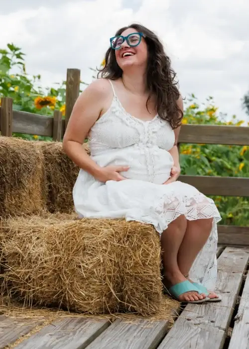image (55) A pregnant woman in a white lace dress sits on hay bales, surrounded by green foliage and sunflower blooms under a cloudy sky.